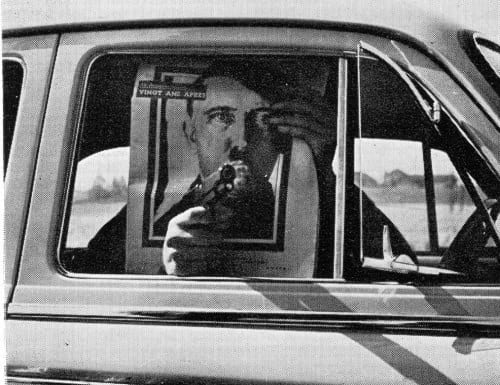 Un tirador apunta su revolver desde un coche tapándose con un cartel de Hitler