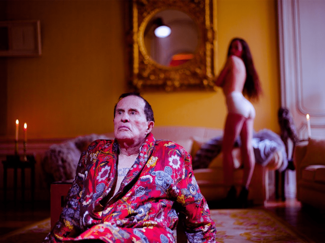 El director posa con un batín muy llamativo y una modelo desnuda y un sofá al fondo