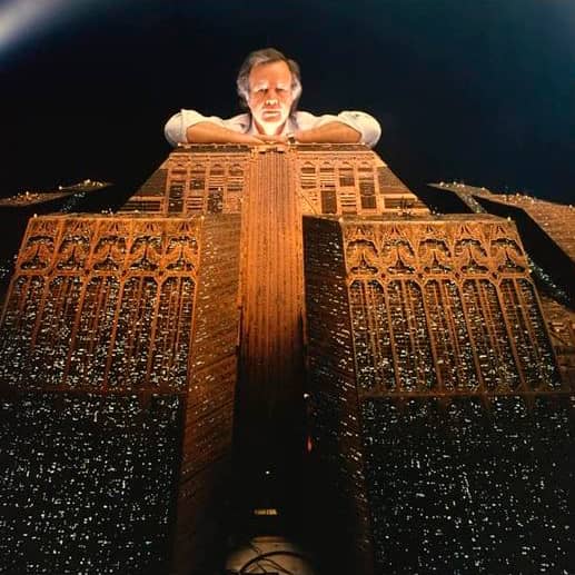 El artista apoyado sobre la gran maqueta de la corporación que manda en el film