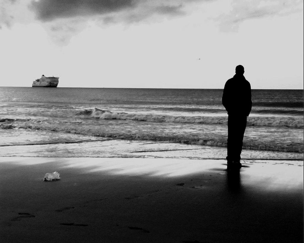 Un inmigrante mira al mar tormentoso donde pasa un barco a lo lejos, en blanco y negro