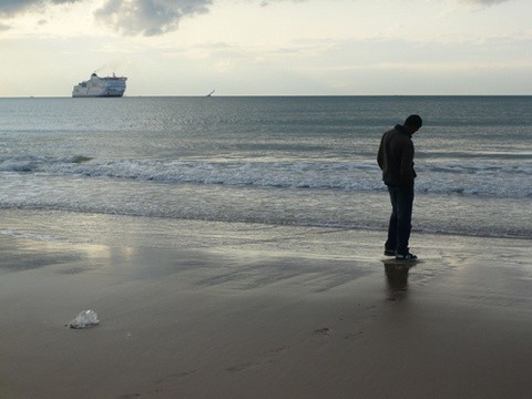 Un inmigrante mira al mar tormentoso donde pasa un barco a lo lejos, en color